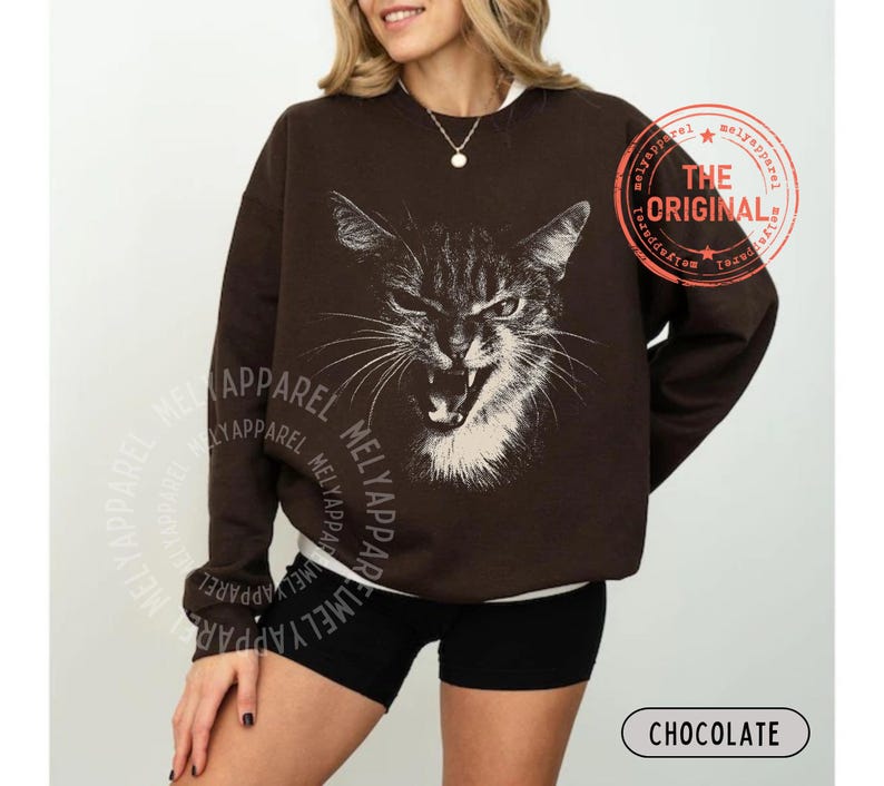 May include: A dark brown long-sleeve sweatshirt featuring a white graphic illustration of a cat with an open mouth and bared teeth. The text "THE ORIGINAL" is printed in a circular red stamp design on the upper right side of the garment.