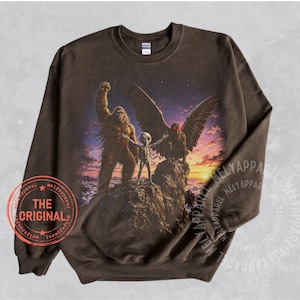 May include: A dark brown crewneck sweatshirt featuring a graphic print of Bigfoot, a grey alien, and a winged creature with red eyes standing on a rocky mountain peak at sunset. A circular red stamp on the lower left reads THE ORIGINAL.