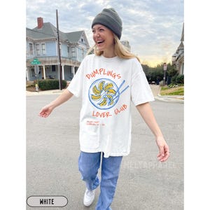 Op de afbeelding: Wit T-shirt met een afbeelding met de tekst "Dumplings Lover Club" in rood en blauw. De afbeelding toont een bord met dumplings en eetstokjes. Het shirt heeft ook de tekst "Friday Night Starting at 7 PM".