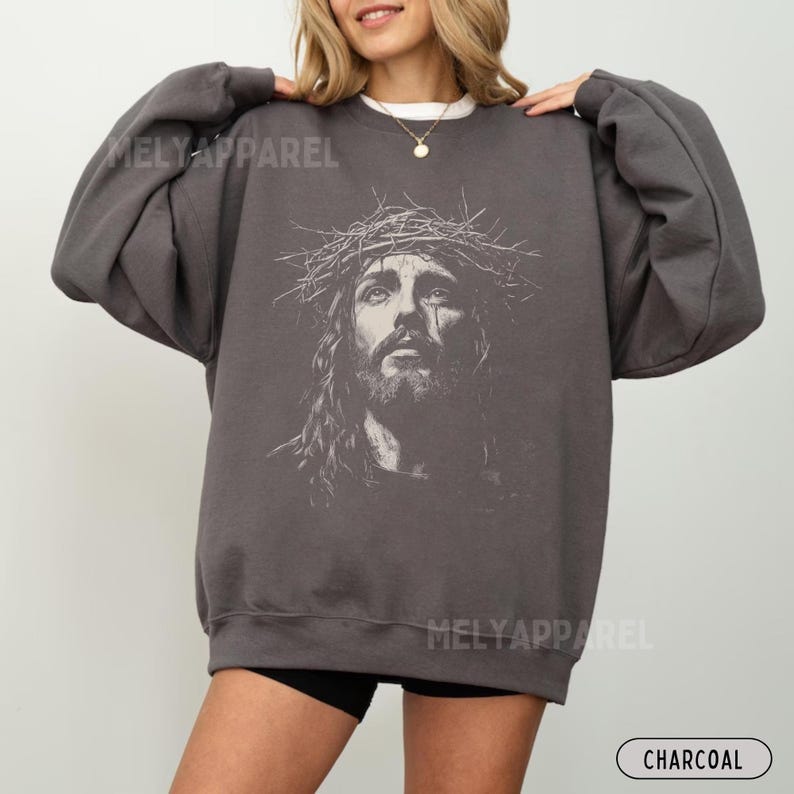 May include: Charcoal-colored sweatshirt featuring a detailed grayscale image of Jesus wearing a crown of thorns. The sweatshirt has long sleeves and the text "MELY APPAREL" is printed on the front. The word "CHARCOAL" is printed at the bottom.