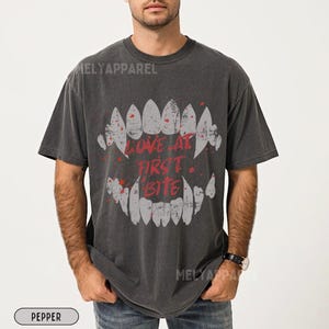 K&ouml;nnte beinhalten: Dunkelgraues T-Shirt mit einem grafischen Motiv aus stilisierten Z&auml;hnen und dem roten Schriftzug "LOVE AT FIRST BITE". Das Design hat einen Used-Look und wirkt vintage.