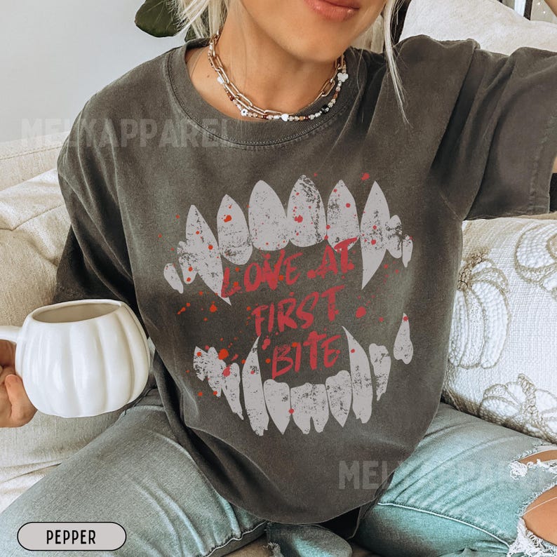 K&ouml;nnte beinhalten: Dunkelgraues T-Shirt mit einer Grafik eines Vampir-Mauls und dem roten Text "LOVE AT FIRST BITE". Das Shirt hat eine lockere Passform und einen Vintage-Look. Eine wei&szlig;e, k&uuml;rbisf&ouml;rmige Tasse wird in der linken Hand gehalten.