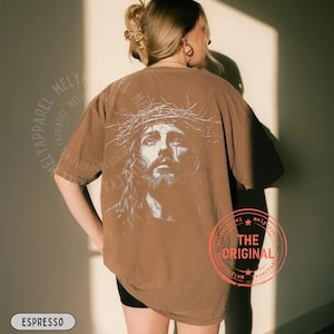 May include: Espresso-colored t-shirt featuring a detailed graphic of a man wearing a crown of thorns. The design is on the back of the shirt. The text "MELY APPAREL" is printed around the shirt's edges. The text "THE ORIGINAL" is printed on a red circle.
