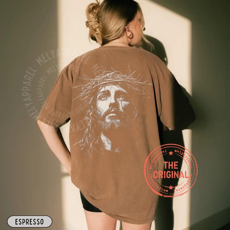 May include: Espresso-colored t-shirt featuring a detailed graphic of a man wearing a crown of thorns. The design is on the back of the shirt. The text "MELY APPAREL" is printed around the shirt's edges. The text "THE ORIGINAL" is printed on a red circle.