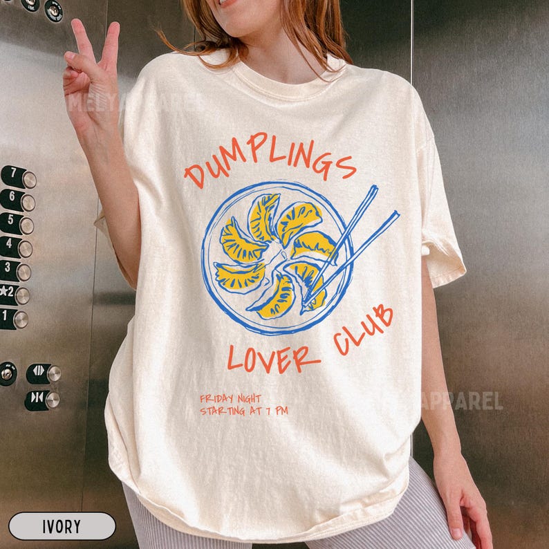 Op de afbeelding: Ivoor t-shirt met een afbeelding van dumplings in een blauwe cirkel met eetstokjes. De tekst "DUMPLINGS LOVER CLUB" staat in het rood, met "FRIDAY NIGHT STARTING AT 7 PM" eronder. Het shirt heeft een losse pasvorm.