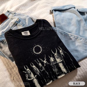 May include: Black t-shirt featuring a graphic print of a group of figures with antlers and a sun with a black circle in the centre. The text "COMFORT COLORS" is visible on the shirt.