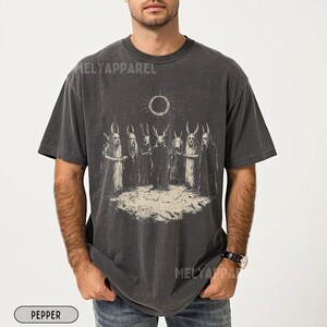 May include: A dark grey t-shirt featuring a graphic print of a group of figures with horns and a sun with a black circle in the centre. The figures are standing in a circle around the sun. The text "MELYAPPAREL" is visible on the shirt.