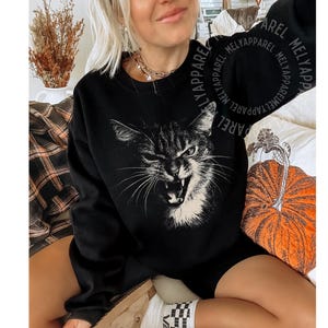May include: A black long-sleeve sweatshirt featuring a large, white, high-contrast graphic print of a cat with an open mouth and bared teeth. The sweatshirt has a crew neckline and long sleeves, with the text MELYAPPAREL repeated in a circular pattern.