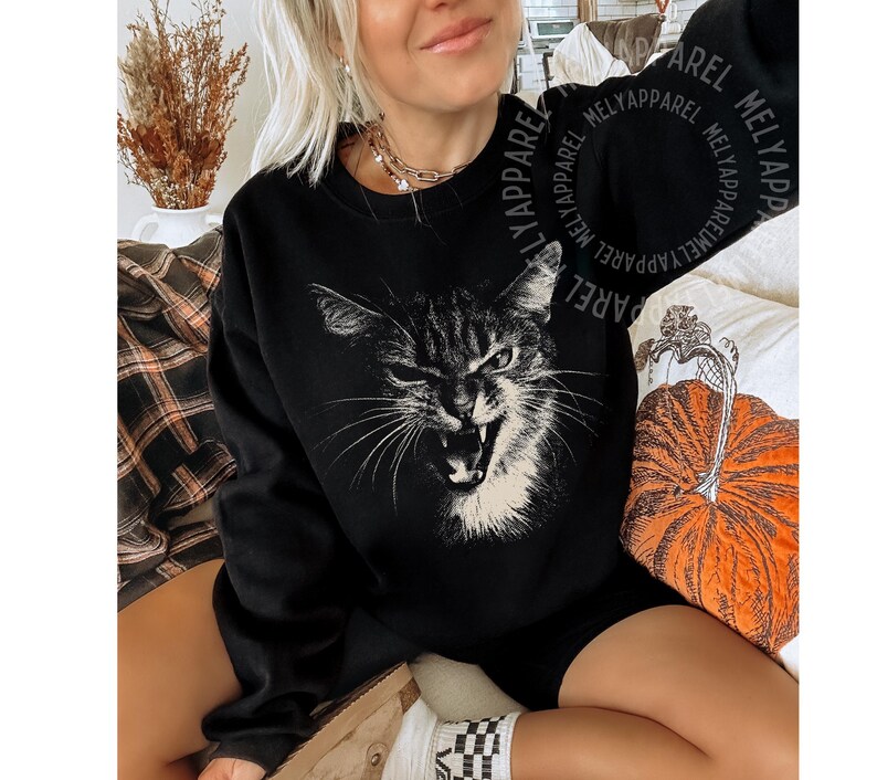 May include: A black long-sleeve sweatshirt featuring a large, white, high-contrast graphic print of a cat with an open mouth and bared teeth. The sweatshirt has a crew neckline and long sleeves, with the text MELYAPPAREL repeated in a circular pattern.