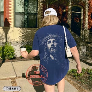 May include: True navy blue t-shirt featuring a detailed illustration of Jesus wearing a crown of thorns. The back of the shirt has the text "Mely Apparel" and "The Original" in a circular stamp. The shirt is a casual wear item.