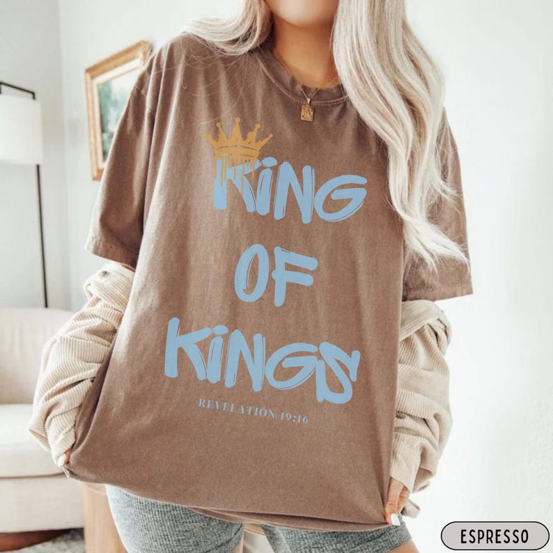 May include: A brown t-shirt with a blue graphic that says "King of Kings" and a gold crown above the text. The text "Revelation 19:16" is printed below the graphic. The t-shirt is being worn by a person.
