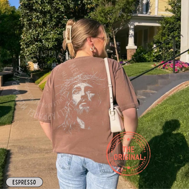 May include: Espresso-colored t-shirt featuring a detailed graphic of a person with a crown of thorns. The shirt has the text "Jewely Apparel Me" and "The Original" printed on it. The model is wearing blue jeans and a white purse.