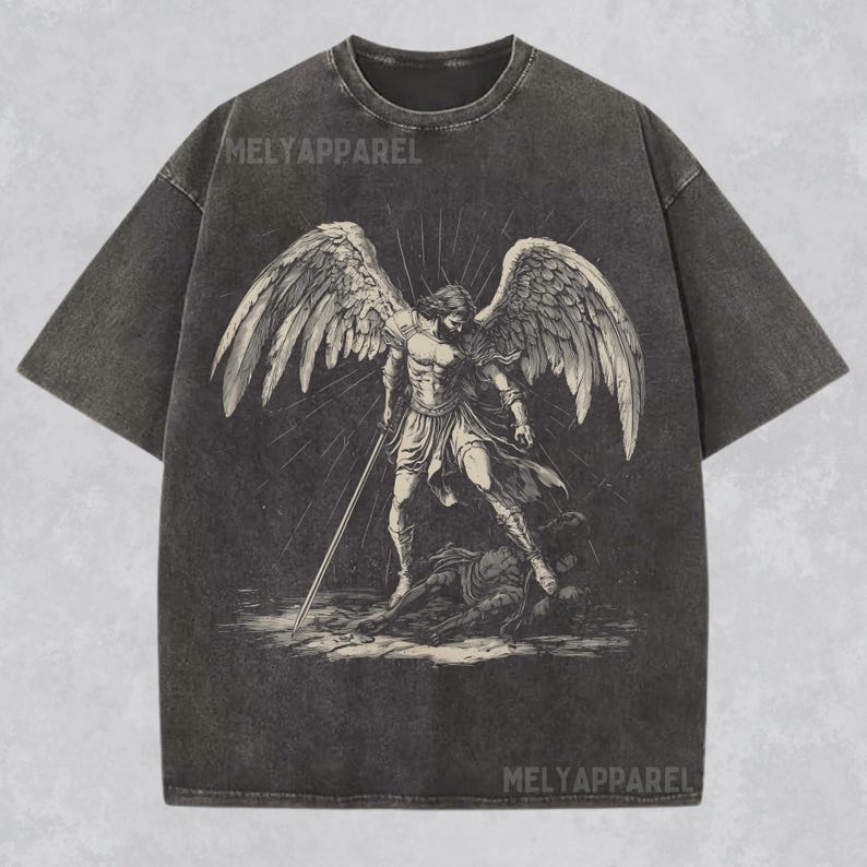 May include: Dark grey t-shirt with a detailed illustration of an angel with large wings, holding a sword, and standing over a defeated figure. The text "MELYAPPAREL" is printed at the top and bottom of the shirt.