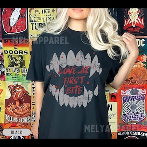 K&ouml;nnte beinhalten: Schwarzes T-Shirt mit einem Zahnmotiv und dem roten Schriftzug "LOVE AT FIRST BITE". Dieses Kleidungsst&uuml;ck ist f&uuml;r den l&auml;ssigen Stil geeignet und ein auff&auml;lliges Statement.