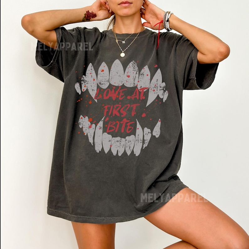 K&ouml;nnte beinhalten: Dunkelgraues T-Shirt mit einem Grafikdesign eines Vampir-Mauls mit spitzen Z&auml;hnen und dem roten Text "LOVE AT FIRST BITE". Das Shirt hat eine lockere Passform und einen Vintage-Look.