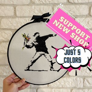 May include: A white embroidery hoop with a black frame, featuring a cross-stitch design of a person holding a flower. The text "Support New Shop" and "Just 5 Colors" is visible in the image.