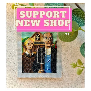 May include: A cross-stitch artwork of a painting depicting three people standing in front of a house. The artwork is framed in white and has a pink background with the text "SUPPORT NEW SHOP" in white.