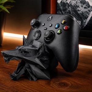 May include: A black gaming controller resting on a matte black, geometric, low-poly wolf head stand. The controller features a central Xbox logo, two analogue sticks, a directional pad, and four coloured action buttons labelled A, B, X, and Y.