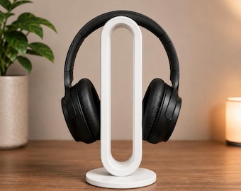 May include: A white headphone stand with a rounded design holds a pair of black over-ear headphones. The stand has a circular base and a vertical support with a curved top. The headphones are positioned on the stand, ready for use.