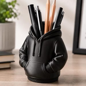May include: A black desk tidy in the shape of a hoodie, filled with pens and pencils. The organiser has a hood, a front pocket, and arm-shaped sides with pockets. The pencils are light brown, and the pens are black and silver.