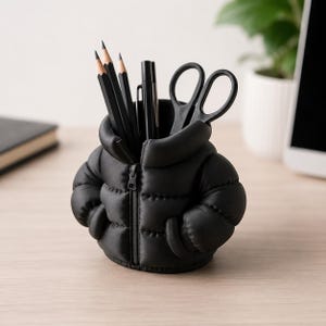 Może przedstawiać: Czarny organizer na biurko w kształcie miniaturowej kurtki puchowej z centralnym zamkiem błyskawicznym. Pojemnik mieści trzy czarne ołówki, czarny długopis oraz czarne nożyczki, stanowiąc funkcjonalne rozwiązanie do przechowywania przyborów.
