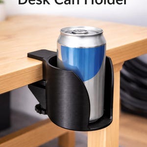 Puede incluir: Un soporte para latas de escritorio negro impreso en 3D, diseñado para sujetarse a una mesa. El soporte sujeta de forma segura una lata plateada con una etiqueta azul y blanca. El texto "3D Printed Desk Can Holder" es visible.