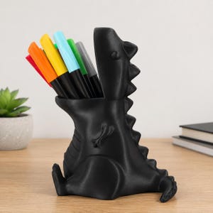 May include: A matte black dinosaur-shaped desk organiser with an open mouth design. The hollow interior holds a collection of felt-tip pens in vibrant colours including red, orange, yellow, light blue, green, and grey, standing upright.