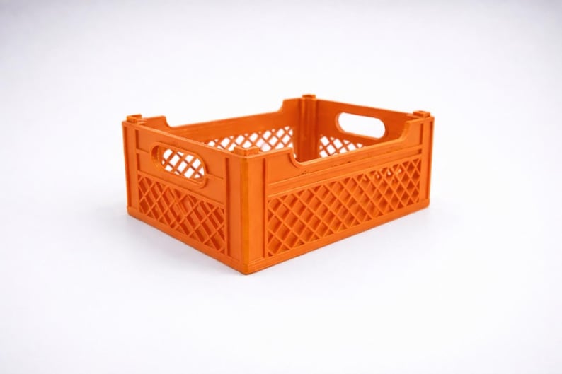 May include: An orange plastic crate. The crate has a rectangular shape with a diamond-patterned design on the sides and handle cutouts. It is empty and sits on a white surface. The crate is likely used for storage.