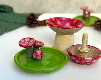 May include: A collection of whimsical ceramic mushroom-shaped objects. The set includes a small green dish with red-capped mushrooms, a larger mushroom-shaped bowl, and a small bowl with a mushroom-like pestle. The mushrooms have red and white speckled caps.