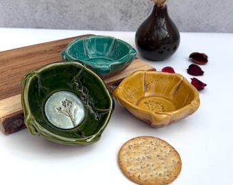 May include: Three ceramic bowls in green, teal, and yellow, each with a unique leaf-like design. The green bowl features a floral imprint. A brown vase with dried flowers and a cracker are also present.