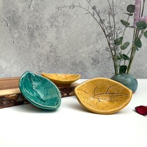 Puede incluir: Tres cuencos de cerámica con forma de hoja en verde azulado, amarillo y dorado. Los cuencos tienen una superficie texturizada y agrietada. Una tabla de madera y un jarrón con flores están en el fondo. Cuencos decorativos para servir aperitivos o exhibir objetos pequeños.