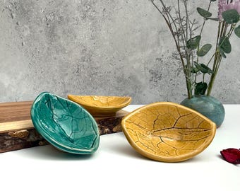 May include: Three ceramic leaf-shaped dishes in teal, yellow, and gold. The dishes have a textured, cracked surface. A wooden board and a vase with flowers are in the background. Decorative bowls for serving snacks or displaying small items.
