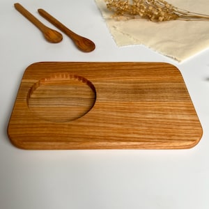 May include: A rectangular wooden serving tray with a circular cutout in the centre. The tray is made of light brown wood with a natural grain pattern.