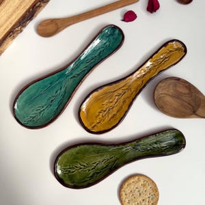 May include: Three ceramic spoon rests in green and yellow with a brown rim and a wheat stalk design. Two wooden spoons are also visible. The spoon rests are on a white surface.