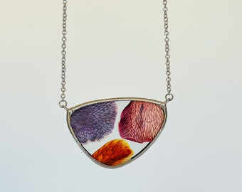 May include: A silver-toned necklace featuring a pendant with a clear, triangular frame. Inside the frame are three colorful, petal-like shapes in shades of purple, red, and orange. The chain is delicate and hangs against a white background.