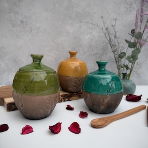 May include: Three ceramic jars with lids, each with a different coloured glaze. The jars are green, yellow, and blue. The jars have a textured, rustic look and are decorated with a leaf pattern. The jars are sitting on a wooden surface.