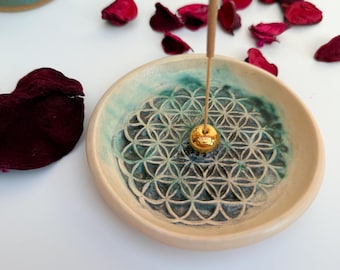 May include: A white ceramic incense holder with a green and blue geometric design. The holder has a small gold ball in the center and a brown incense stick is placed in the holder.