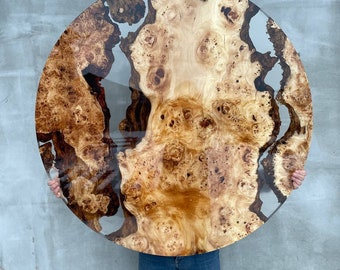 May include: A large round wooden table top with a natural wood grain pattern and clear epoxy resin filling in the gaps. The wood is a light brown color with dark brown streaks and knots. The epoxy resin is clear and shiny.