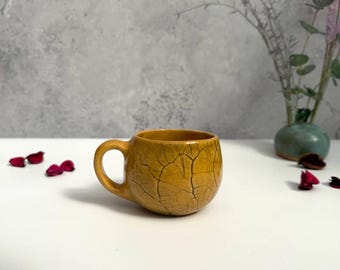 May include: A small, round, mustard-yellow ceramic mug with a single handle. The mug has a textured, cracked surface. The background includes a gray wall, a small vase with flowers, and scattered red petals.
