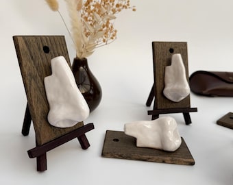 May include: White ceramic nose sculptures displayed on small wooden easels and a wooden base. The sculptures are set against a neutral background with a vase of dried flowers. The wooden easels and base have a dark brown finish.