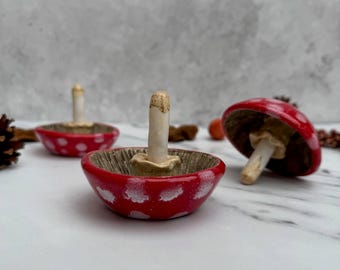 May include: Ceramic mushroom-shaped ring holders. Each holder features a red cap with white polka dots and a beige stem. The interior of the bowl has a textured, wood-like appearance. One mushroom is upside down, revealing the underside.