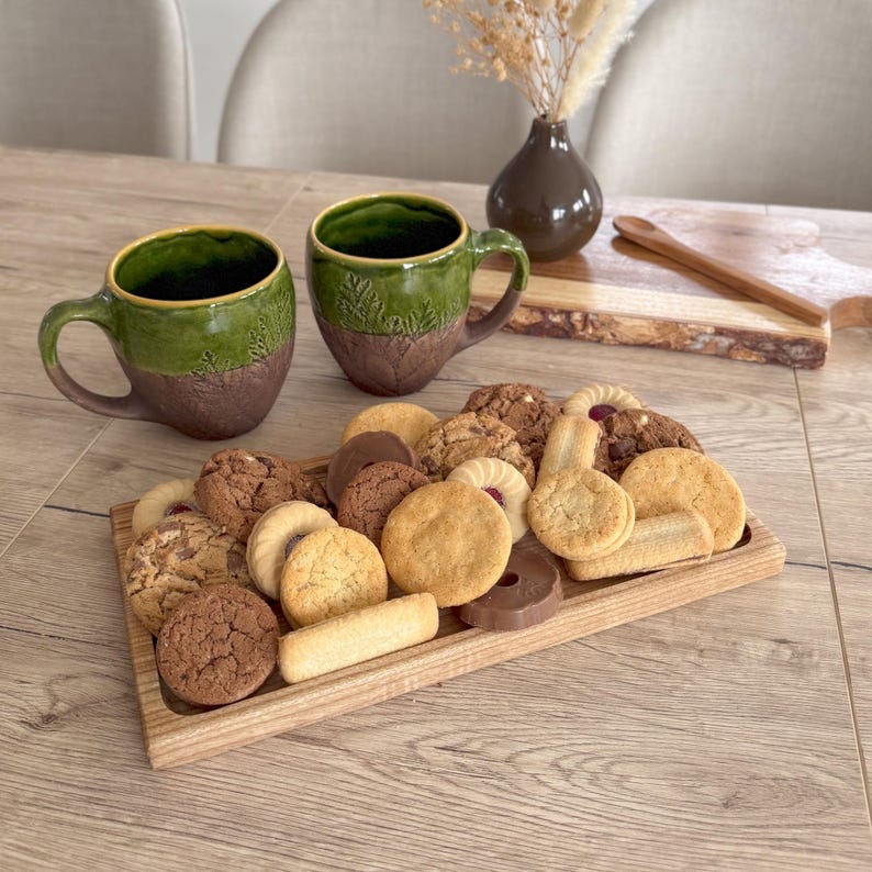May include: A wooden tray filled with a variety of cookies, including shortbread, chocolate chip, and biscotti. Two green ceramic mugs with brown accents are on the table next to the tray.