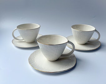 May include: Three white ceramic teacups with gold rims and matching saucers. The cups and saucers have a textured surface.
