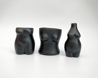 May include: Three black ceramic vases shaped like the human body. The vases are all different sizes and shapes, with one being a more traditional vase shape, one being a more hourglass shape, and one being a more rounded shape.