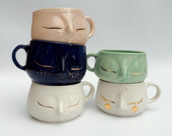 May include: Four ceramic mugs with a cat face design. The mugs are stacked in two pairs. The top two mugs are pink and blue, the bottom two are white and speckled white. All mugs have gold painted eyes and a gold painted nose.