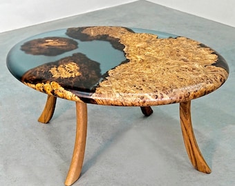 May include: A round wooden coffee table with a unique epoxy resin inlay. The table has a light blue and dark brown epoxy resin design and four curved wooden legs.