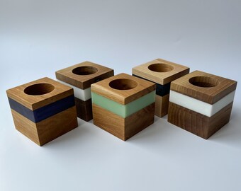 May include: Four wooden square candle holders with different colored resin inlays. The holders are made of natural wood with a light brown finish. The resin inlays are blue, green, white, and black.