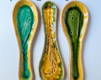 May include: Three ceramic spoon rests with green and yellow glaze. Each rest features a different design of stylized green and yellow foliage.