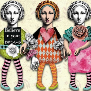 Puede incluir: Tres muñecas de papel con ropa y accesorios coloridos. La muñeca de la izquierda lleva un vestido verde y blanco con las palabras "Cree en tus sueños" en un cartel negro. La muñeca del medio lleva un corazón rojo y un vestido con estampado de diamantes. La muñeca de la derecha lleva un vestido rosa con una flor grande.