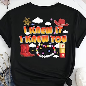 May include: A black t-shirt featuring a graphic print with the text I KNEW IT I KNEW YOU. The design includes a red cowboy hat, a sheriff star, red cowboy boots, a beaded necklace with the word BESTIES, dice, and three stacked blocks with TSS.